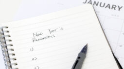 A pen rests on a notebook where someone has written “New Year’s Resolutions” and a blank list numbered one through three.